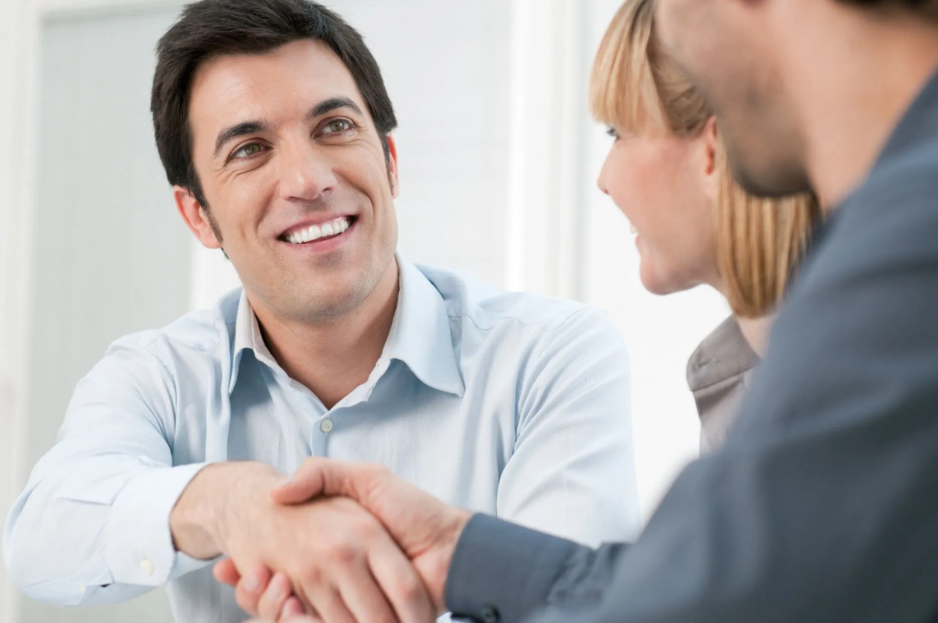 A man smiling and shaking hands with another man in an office while a woman watches.
