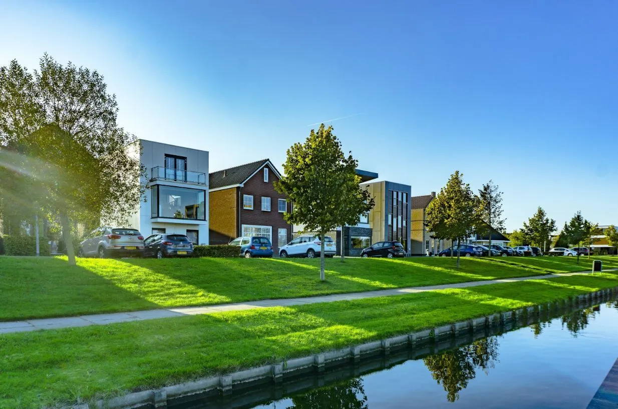 Houses line a canal on a sunny day, with green grass and trees in the foreground under a blue sky.