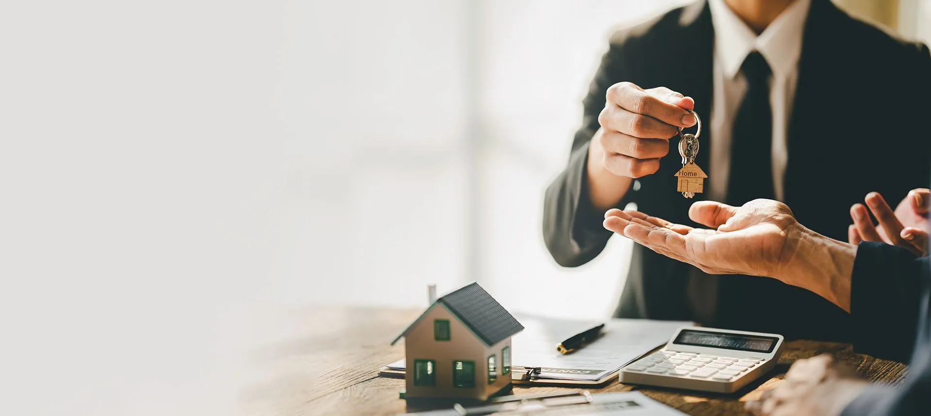 A property manager in a suit hands over house keys with a wooden Home keychain, beside a model house and calculator.