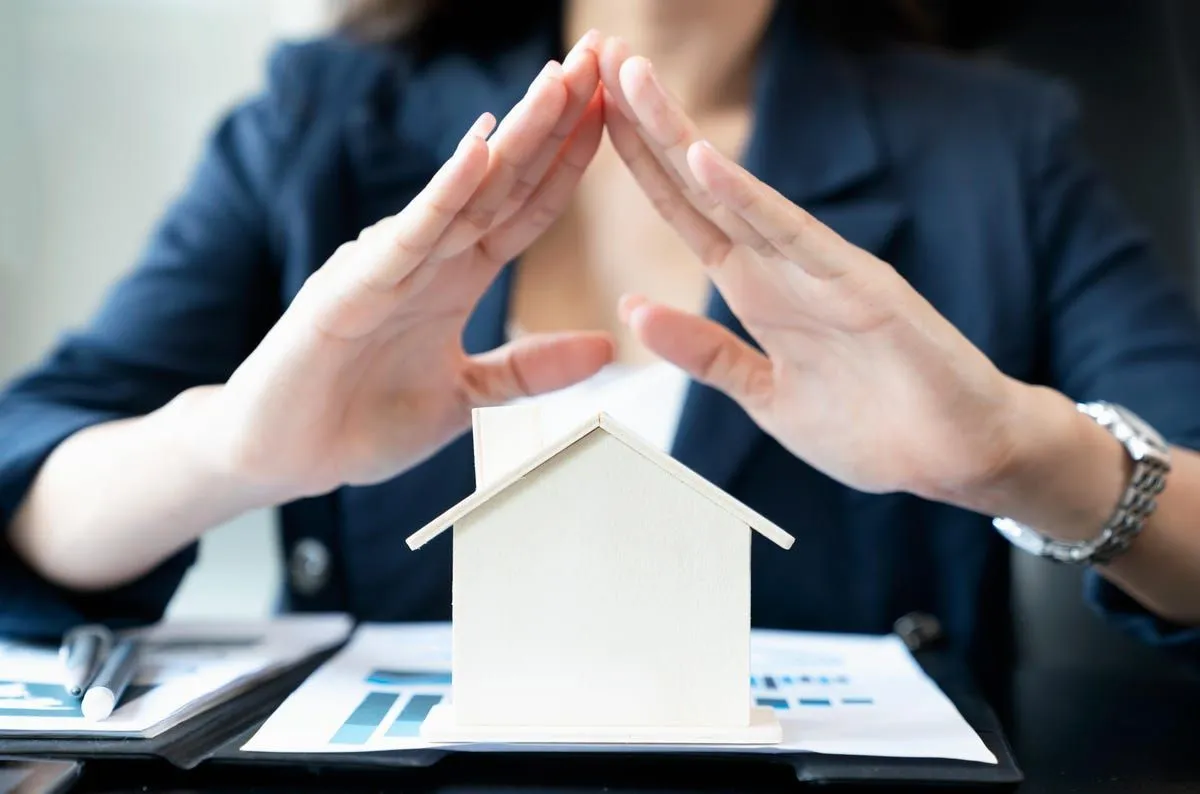 A woman's hands protect a small model house on a desk with papers.