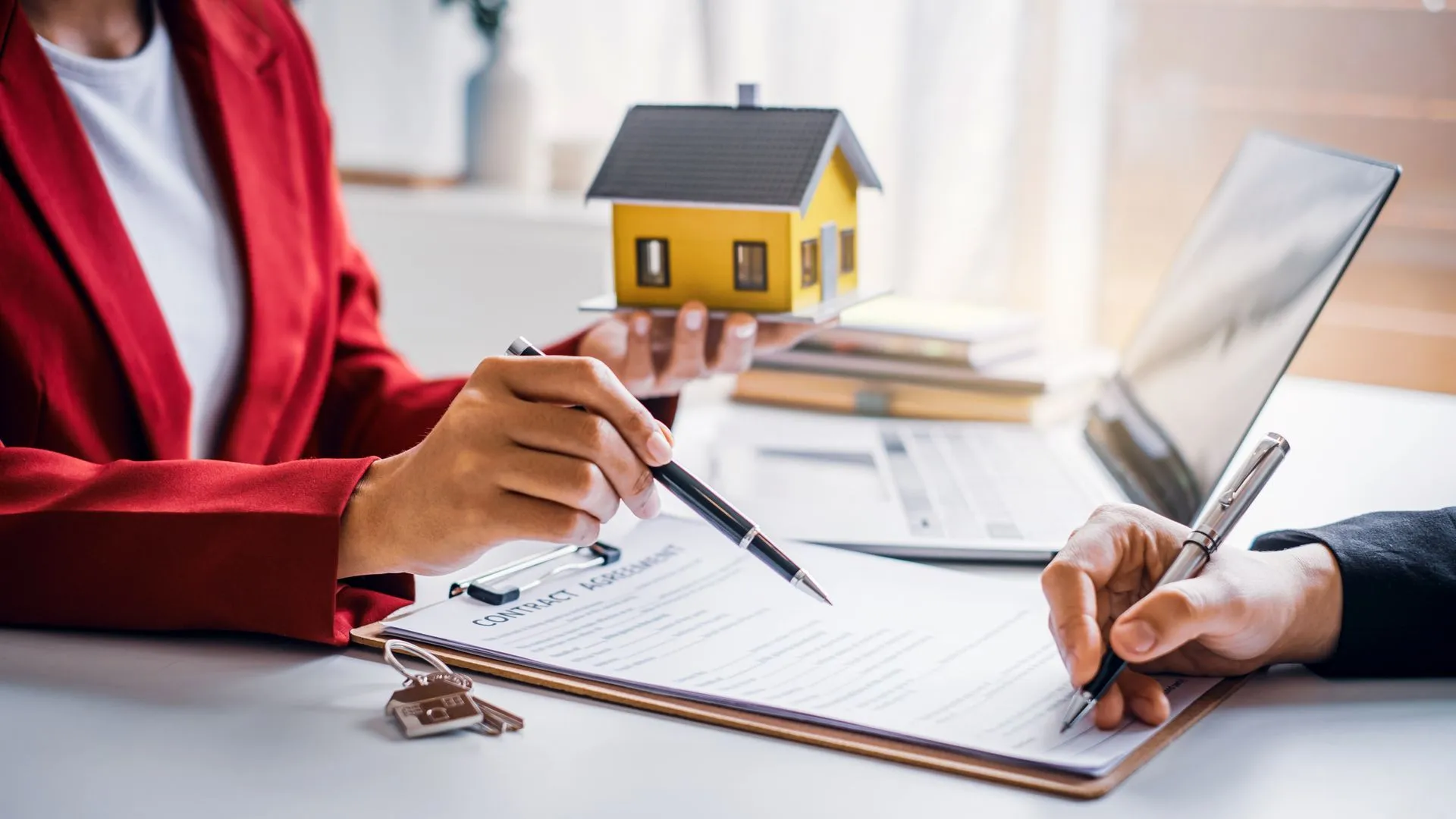 A woman in a red jacket points at paperwork as another person signs, with a house model present.