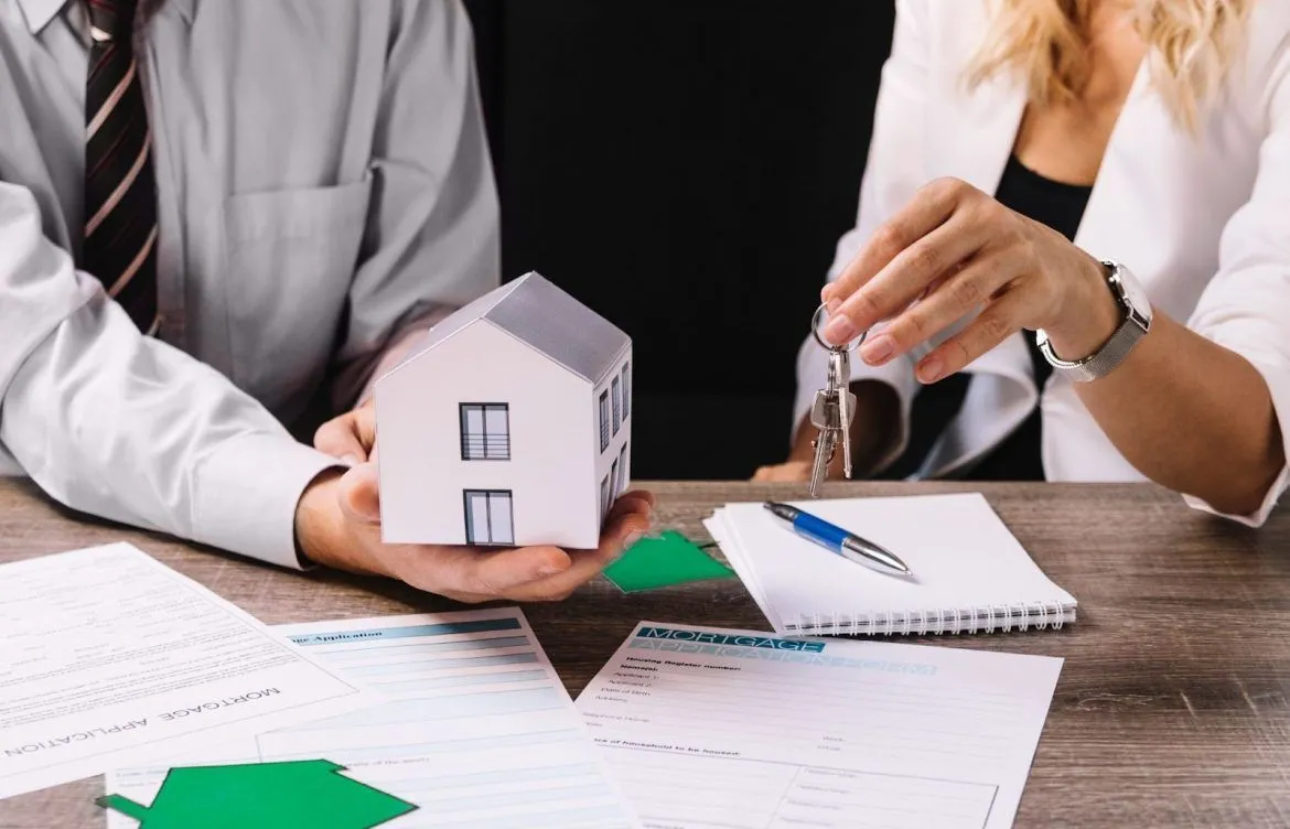 A person holding a model house while another holds keys, with documents on a desk.
