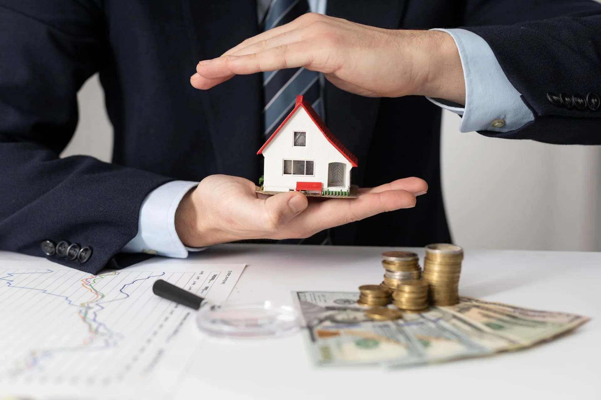 A person in a suit protects a model house with their hands, near money and coins on a desk.
